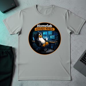 Puede incluir: Camiseta gris claro con un diseño circular. El diseño muestra un pingüino de dibujos animados sentado frente a un ordenador, sosteniendo una taza. El texto en el diseño dice "Homelab HONORED MEMBER" en blanco y naranja.