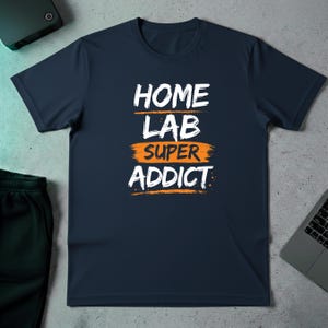 以下が含まれることがあります： 白とオレンジ色の「HOME LAB SUPER ADDICT」の文字がプリントされたネイビーブルーのTシャツ。テキストは筆のストローク効果で3行に配置されています。Tシャツは明るい灰色の表面にあります。