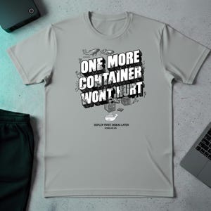 May include: A light gray t-shirt with the text "ONE MORE CONTAINER WON'T HURT" in a 3D block font. The shirt also features small illustrations of whales, containers, and a ship. Below the text is "DEPLOY FIRST, DEBUG LATER".