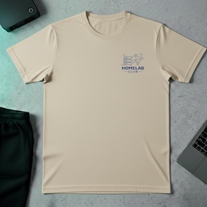 Puede incluir: Una camiseta beige de cuello redondo con un pequeño gráfico azul y el texto "HOMELAB CLUB" en la parte superior izquierda del pecho. La camiseta está hecha de un material ligero y tiene mangas cortas. La camiseta está sobre una superficie texturizada.