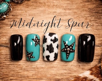 May include: A set of five artificial nails with a western theme. The nails are black, turquoise, and cow print, with star accents. The text "Midnight Spur" is written in cursive. The background is wood.