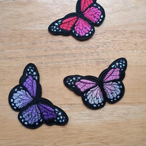 May include: Three embroidered butterfly patches in shades of pink, purple, and white, with black outlines and white dot details. The patches are arranged on a light brown wooden surface.