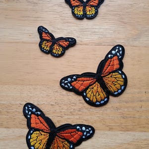 May include: Four embroidered butterfly patches in various sizes. The butterflies have orange and yellow wings with black outlines and details. The patches are on a light brown wooden surface.