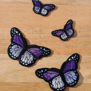 May include: Four embroidered butterfly patches in different sizes. The butterflies have purple wings with white and black detailing. The patches are displayed on a light brown wooden surface.