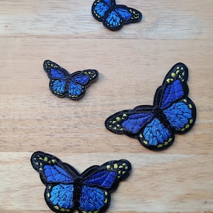 May include: Four embroidered butterfly patches in different sizes. The butterflies are a vibrant blue with yellow and black detailing. They are displayed on a light brown wooden surface.