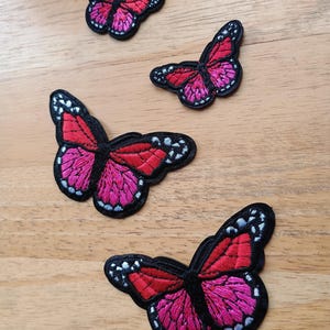 May include: Four embroidered butterfly patches in varying sizes. The butterflies have red and pink wings with black outlines and white and blue accents. The patches are arranged on a light brown wooden surface.