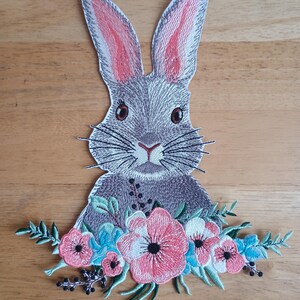 May include: Embroidered patch of a grey bunny with pink inner ears and brown eyes. The bunny is decorated with a floral arrangement of pink, blue, and white flowers and green leaves, ideal for customising clothing or accessories.