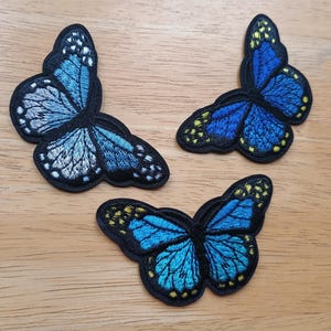 May include: Three embroidered butterfly patches in shades of blue, black, and yellow. The butterfly wings have detailed stitching and are set against a black border. The patches are arranged on a light brown wooden surface.
