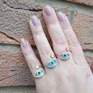 May include: A hand showing three adjustable eye-shaped rings. The rings have a turquoise centre, encircled by small, clear stones. One ring is gold-toned, one is silver-toned, and one is rose gold-toned.