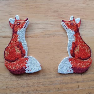 May include: Two embroidered fox patches. Each fox is depicted in a sitting position, with its head tilted upwards. The foxes have red and white fur, with detailed stitching. The patches are likely for clothing or craft projects.