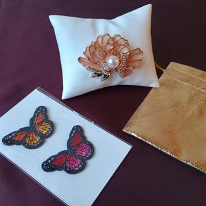 May include: A white cushion with a peach flower brooch, pearl centre, and rhinestone details. Two butterfly patches, one orange and yellow, the other red and pink, are on a white card. A gold pouch is also present.