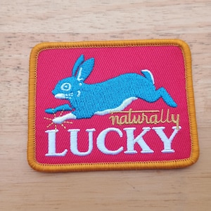May include: Rectangular embroidered patch featuring a blue rabbit in motion on a pink backdrop. The patch is edged with gold thread and displays the words "naturally LUCKY" in white and gold.