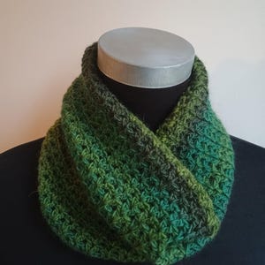 May include: A green and dark green crocheted infinity scarf. The scarf is displayed on a black mannequin and has a textured, handmade appearance. The color gradient transitions from lighter to darker shades of green.