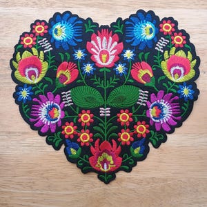 May include: A heart-shaped embroidered patch with a black background, featuring a vibrant floral design. The flowers are in shades of red, pink, blue, yellow, and purple, with green leaves. The patch is likely for decorative purposes.