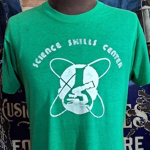 May include: A green t-shirt featuring the white text "SCIENCE SKILLS CENTER" and a graphic. The t-shirt is displayed on a mannequin. The shirt is made of a soft, comfortable material.