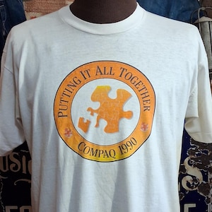May include: A vintage white t-shirt with a crew neck and short sleeves. The shirt features a circular graphic with the text "PUTTING IT ALL TOGETHER COMPAQ 1990" around an orange puzzle piece design. The shirt is displayed on a mannequin.