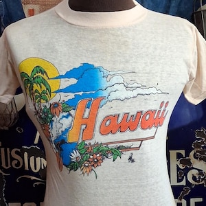 May include: A light beige t-shirt with a graphic of a tropical scene, including a sun, palm trees, and the word "Hawaii" in orange and red. The shirt is displayed on a mannequin.