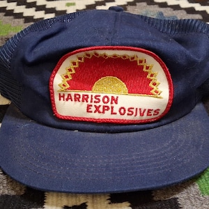Puede incluir: Gorra de camionero azul marino con visera curva. La gorra presenta un parche con las palabras "HARRISON EXPLOSIVES" y un gráfico de un sol naciente. El parche es rojo, blanco y dorado. La gorra está sobre una alfombra estampada.
