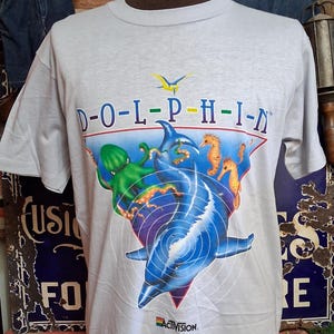 Puede incluir: Una camiseta gris claro con un gráfico colorido de un delfín saltando fuera del agua, con la palabra "D-O-L-P-H-I-N" arriba. El gráfico incluye otras criaturas marinas y el logotipo de Activision.