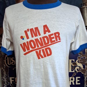 Puede incluir: Camiseta blanca con ribete azul en el cuello y las mangas. La camiseta presenta el texto rojo "I'M A WONDER KID" en una fuente retro audaz. La camiseta se exhibe en un maniquí.