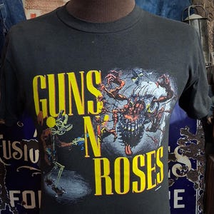 以下が含まれることがあります： 前面に黄色の「GUNS N' ROSES」の文字がプリントされた黒の半袖Tシャツ。グラフィックには、頭蓋骨のような頭部を持つ多肢の生き物の中央イラストと、左側に小さな人物が描かれています。