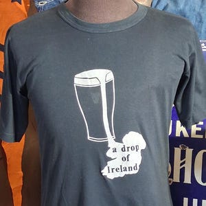 Op de afbeelding: Zwart t-shirt met een witte afbeelding van een pint bier en de omtrek van Ierland, met de tekst "a drop of Ireland". Het t-shirt wordt op een mannequin gepresenteerd.