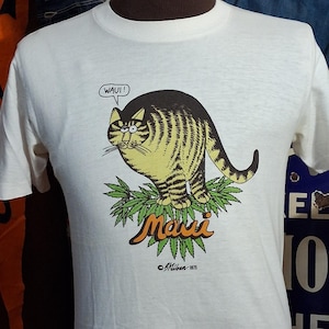 May include: Off-white t-shirt featuring a cartoon cat with yellow and brown stripes, sitting on green leaves. The text "Maui" is above the leaves, and "WAUI!" is in a speech bubble. The shirt is from &copy;Mibon 1975.
