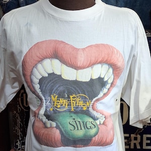 Może przedstawiać: Biała koszulka z grafiką otwartych ust z czerwonymi ustami i białymi zębami. Grafika zawiera tekst "Monty Python Sings" w kolorze żółtym i zielonym. Koszulka jest prezentowana na manekinie.