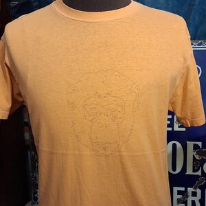 Op de afbeelding: Een oranje t-shirt met korte mouwen en een subtiele, monochrome lijntekening van een chimpanseegezicht in het midden op de borst. Het kledingstuk heeft een klassieke ronde hals en een standaard pasvorm, gemaakt van effen katoenen stof.