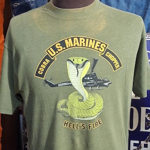 Pode incluir: T-shirt verde-oliva com um gráfico de uma cobra e um helicóptero, com o texto "COBRA U.S. MARINES CHOPPER" e "HELL'S FIRE". A camisa é exibida em um manequim.