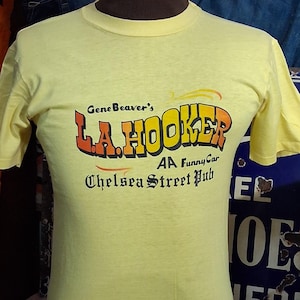 May include: A pale yellow t-shirt featuring the text "Gene Beaver's L.A. Hooker AA Funny Car Chelsea Street Pub" in orange and red. The shirt is displayed on a mannequin.