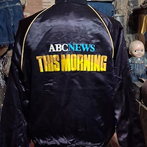 May include: Black satin bomber jacket with yellow trim and the text "ABC NEWS THIS MORNING" in blue and yellow. The jacket has a ribbed collar, cuffs, and waistband. The jacket is displayed on a mannequin.