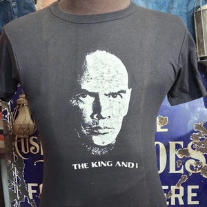 May include: A black short-sleeved t-shirt featuring a white graphic print of a bald man's face centered on the chest. Below the face, the text reads THE KING AND I in white capital letters. The shirt is displayed on a mannequin torso.