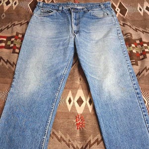 May include: A pair of light blue denim jeans featuring a classic five-pocket design, a metal button closure at the waist, a zippered fly, and visible belt loops. The fabric displays a faded wash pattern across the thighs and knees.