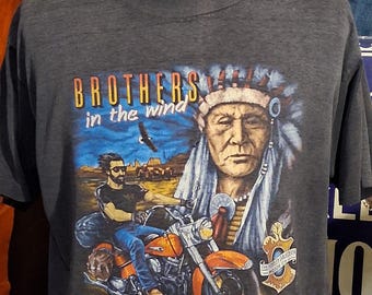 May include: A charcoal gray short-sleeve t-shirt featuring a graphic print with the text BROTHERS in the wind. The design depicts a person riding an orange motorcycle, an eagle, and a portrait of an Indigenous person in a feathered headdress.