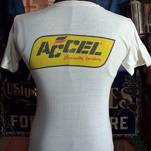 K&ouml;nnte beinhalten: Wei&szlig;es T-Shirt mit gelb-schwarzem ACCEL Eliminator Ignition Logo auf dem R&uuml;cken. Das T-Shirt wird auf einer Schaufensterpuppe pr&auml;sentiert. Das Vintage-T-Shirt hat kurze &Auml;rmel und einen Rundhalsausschnitt. Das Design ist im Retro-Stil.