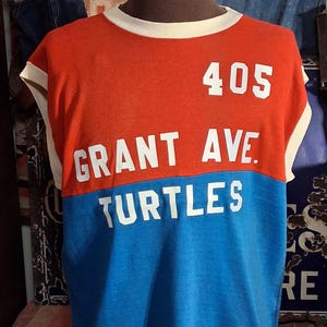 May include: A sleeveless, vintage-style t-shirt in red and blue with white trim. The shirt features the text "405 GRANT AVE. TURTLES" in white block letters. The shirt is displayed on a mannequin.
