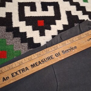 Puede incluir: Una prenda gris oscuro con una regla de madera colocada encima. La regla tiene el texto "An EXTRA MEASURE Of Service" impreso, con marcas de pulgadas visibles. El fondo presenta un textil estampado.