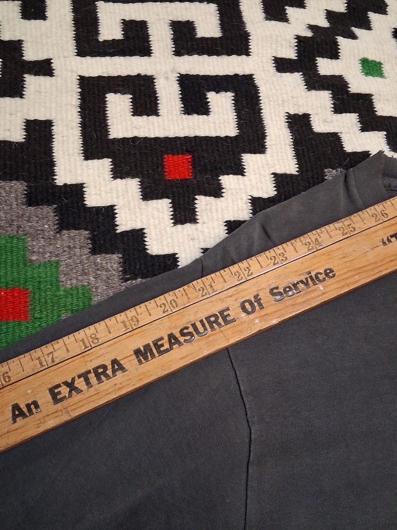 Puede incluir: Una prenda gris oscuro con una regla de madera colocada encima. La regla tiene el texto "An EXTRA MEASURE Of Service" impreso, con marcas de pulgadas visibles. El fondo presenta un textil estampado.