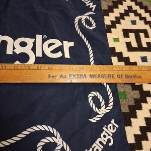 K&ouml;nnte beinhalten: Ein marineblaues Wrangler-Bandana mit wei&szlig;em Seilrand und Logo. Das Bandana ist gefaltet, und ein Holzlineal liegt unten. Der Text "For An EXTRA MEASURE Of Service" ist sichtbar.