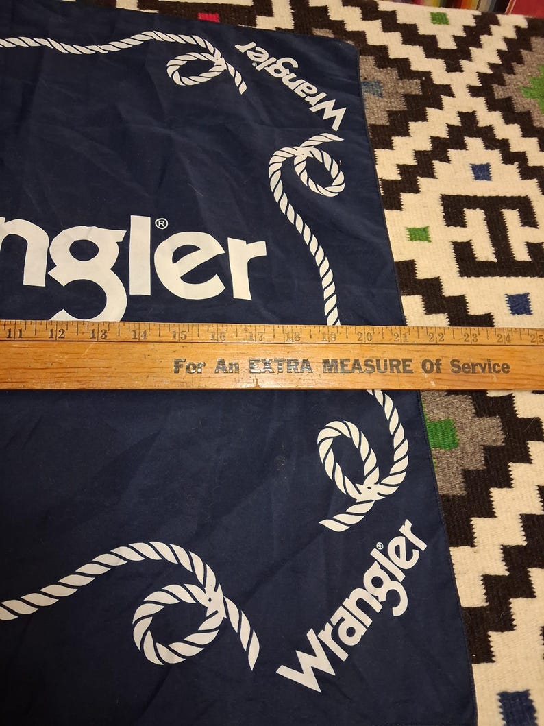 K&ouml;nnte beinhalten: Ein marineblaues Wrangler-Bandana mit wei&szlig;em Seilrand und Logo. Das Bandana ist gefaltet, und ein Holzlineal liegt unten. Der Text "For An EXTRA MEASURE Of Service" ist sichtbar.