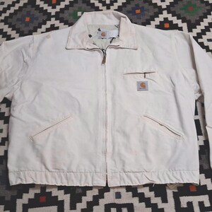 May include: A cream-colored Carhartt zip-up jacket featuring a fold-down collar, a full-length front zipper, two slanted lower pockets, and a single zippered chest pocket with a small rectangular Carhartt brand patch sewn onto the left side.