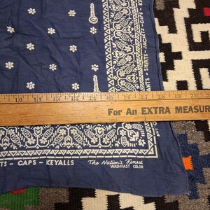 K&ouml;nnte beinhalten: Ein marineblaues Bandana mit wei&szlig;en Blumen- und geometrischen Mustern. Auf dem Bandana steht der Text "ANTS - SHIRTS - JACKETS - CAPS - KEYALLS" und "The Nation's Finest WASHFAST COLOR". Ein Holzlineal liegt darauf.