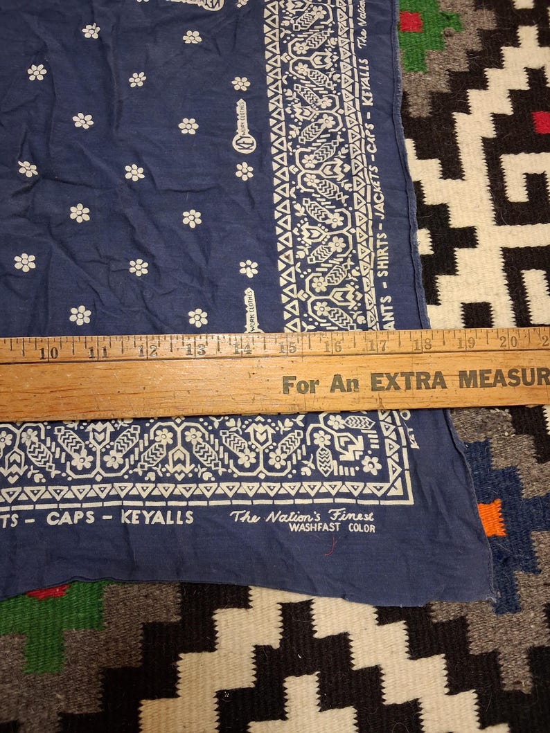 K&ouml;nnte beinhalten: Ein marineblaues Bandana mit wei&szlig;en Blumen- und geometrischen Mustern. Auf dem Bandana steht der Text "ANTS - SHIRTS - JACKETS - CAPS - KEYALLS" und "The Nation's Finest WASHFAST COLOR". Ein Holzlineal liegt darauf.