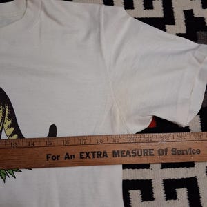 May include: A white t-shirt with a graphic design. A wooden ruler is placed across the shirt. The text on the ruler reads "For An EXTRA MEASURE Of Service".
