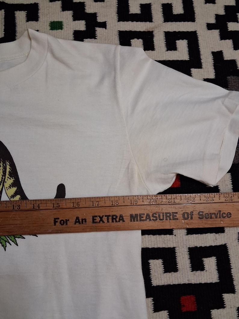May include: A white t-shirt with a graphic design. A wooden ruler is placed across the shirt. The text on the ruler reads "For An EXTRA MEASURE Of Service".
