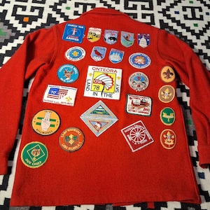 May include: A red long-sleeved jacket adorned with numerous patches. The patches display text, flags, and illustrations of animals and symbols. The jacket has a collar and buttons. One patch reads "ONTEORA 78 IN THE SKY".