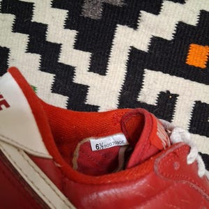 May include: Red and white athletic shoe with a leather upper. The shoe features a white swoosh and laces. The size tag inside the shoe reads "6 1/2 900709DE". The shoe is set against a patterned black and white background.