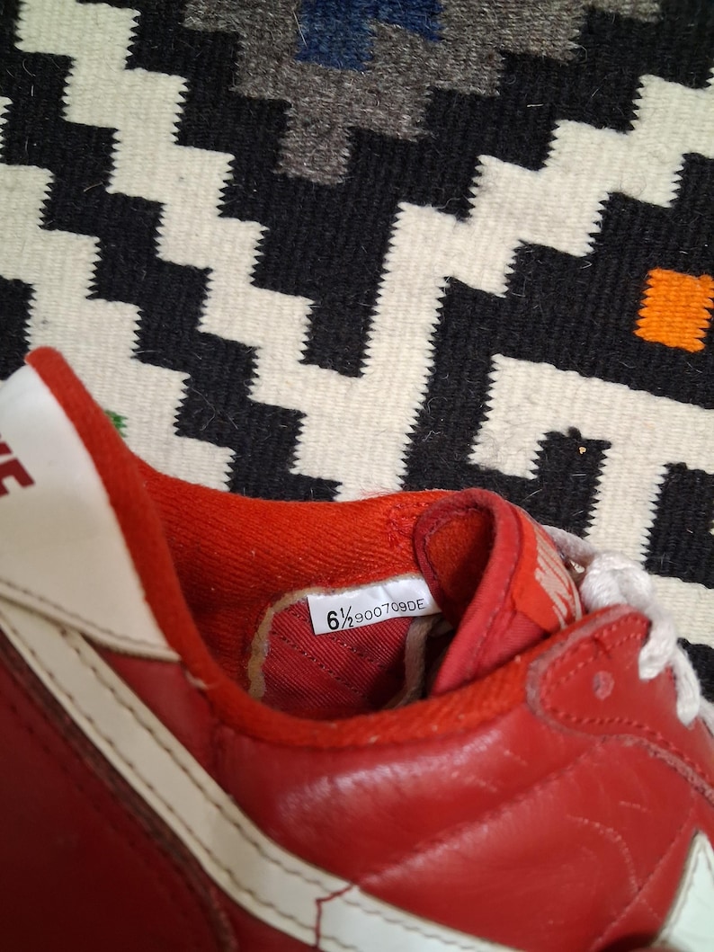 May include: Red and white athletic shoe with a leather upper. The shoe features a white swoosh and laces. The size tag inside the shoe reads "6 1/2 900709DE". The shoe is set against a patterned black and white background.