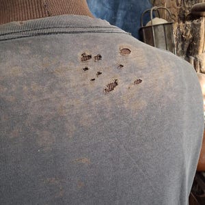 Puede incluir: Una camiseta azul descolorida con varios agujeros en la parte superior de la espalda. La camiseta parece desgastada y tiene una est&eacute;tica vintage. La tela muestra signos de desgaste, con decoloraci&oacute;n y manchas.